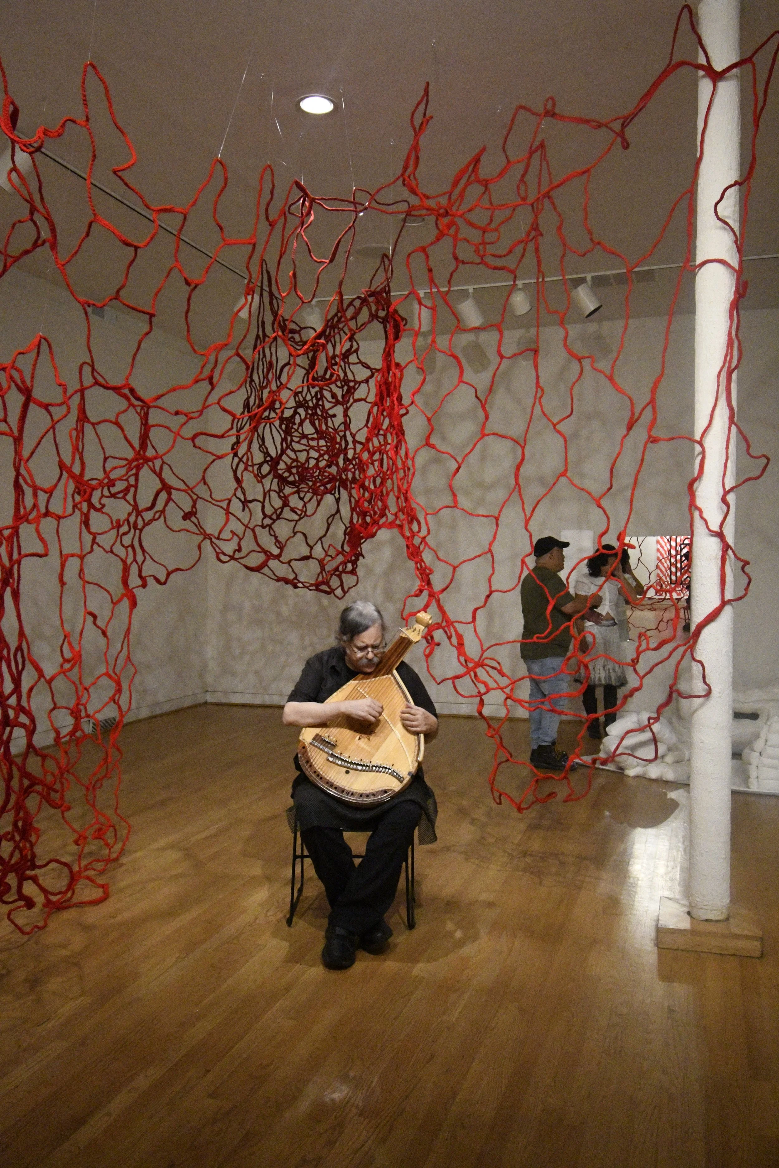 A musician performing on a bandura beneath a large suspended red yarn sculpture installation in the gallery