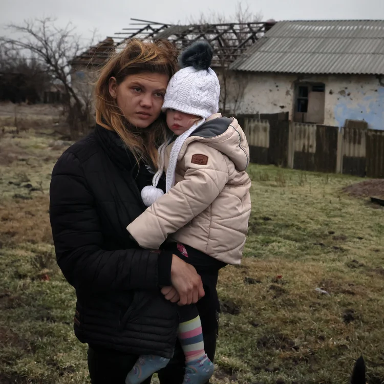 A mother holding her young child outside a war-damaged home in Ukraine