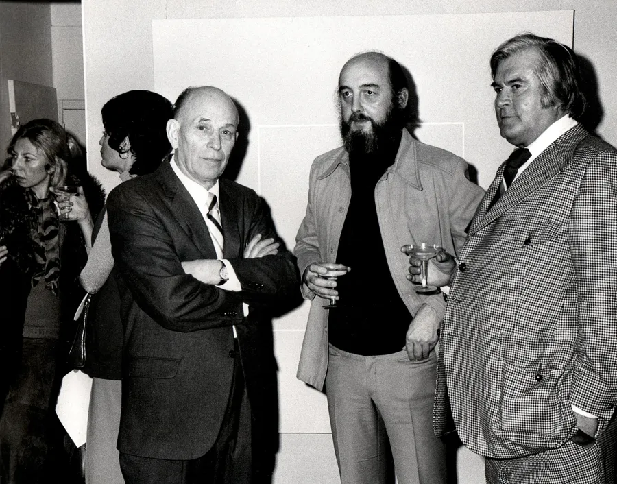Three men in conversation at a gallery reception, each holding a drink