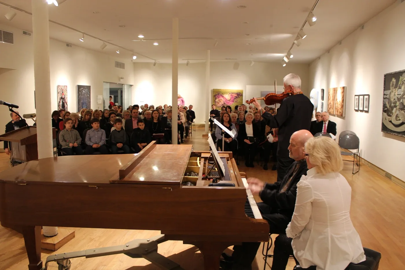 A packed audience at a members' concert in UIMA's gallery, with a violinist performing beside a grand piano