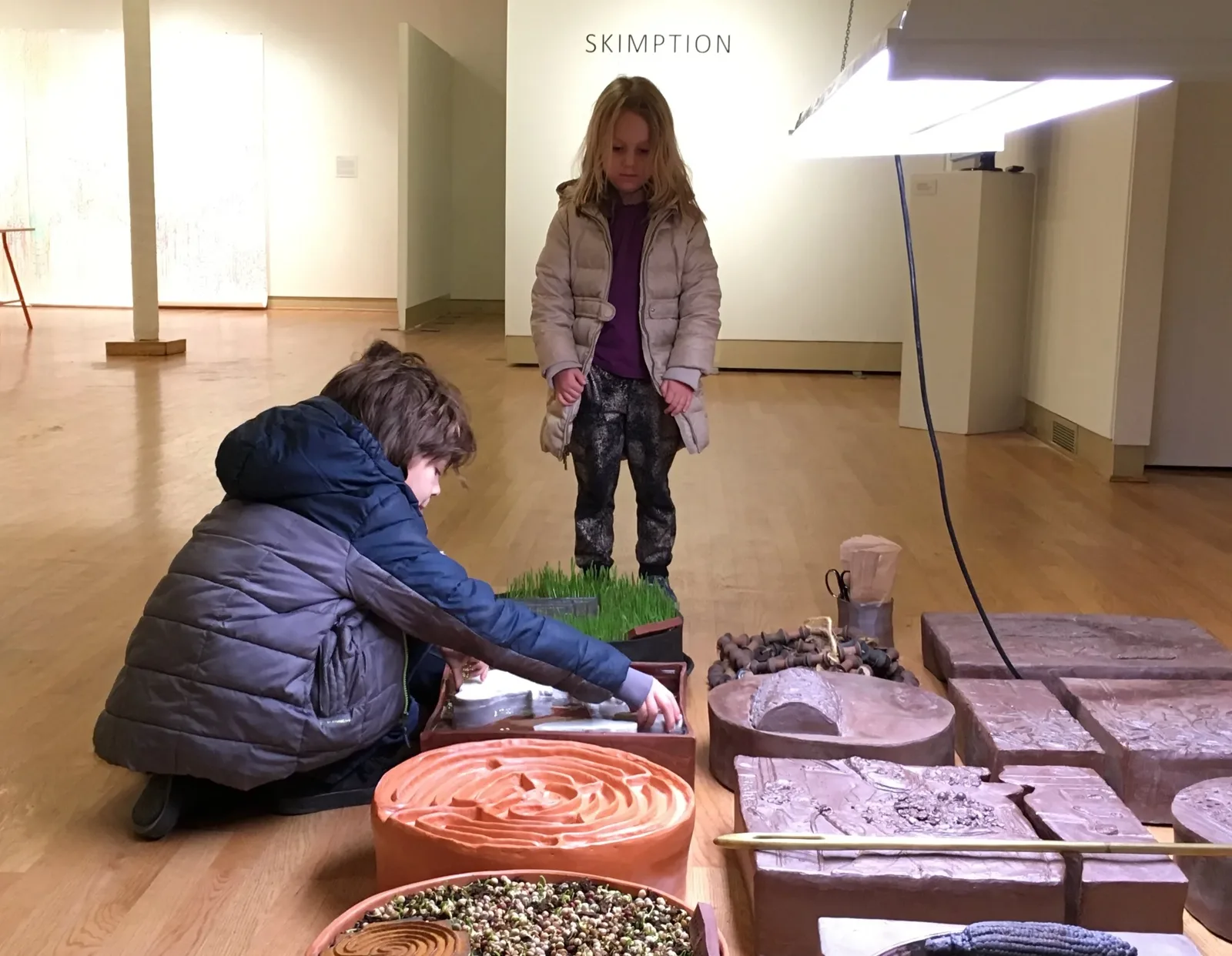 Two children crouched beside a hands-on clay and wheatgrass installation in the gallery